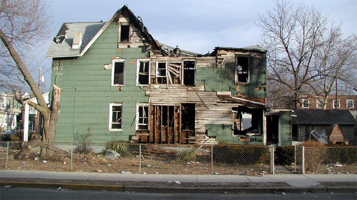 Fire-gutted green clapboard house behind a fence